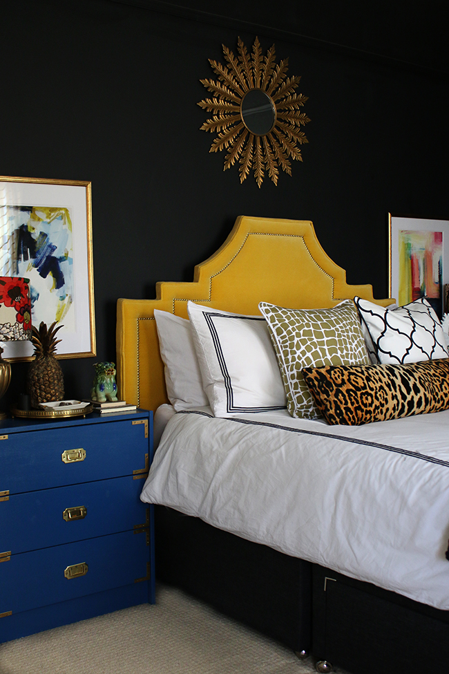 black bedroom with yellow velvet headboard, leopard print cushion, blue rast hack and colourful abstract art - see more on www.swoonworthy.org