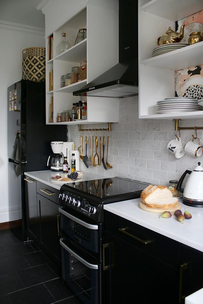 Kitchen with black appliances and cupboards with white open shelving, marble tiles and brass accents