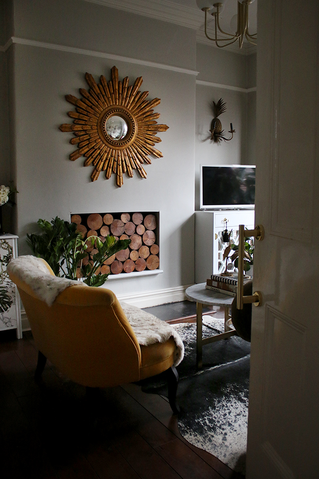 pale grey living room with large gold starburst mirror decorative wood in fireplace and cowhide rug with yellow velvet loveseat